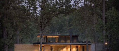 A modern two-story house with a large open patio area, surrounded by a forest and a body of water.