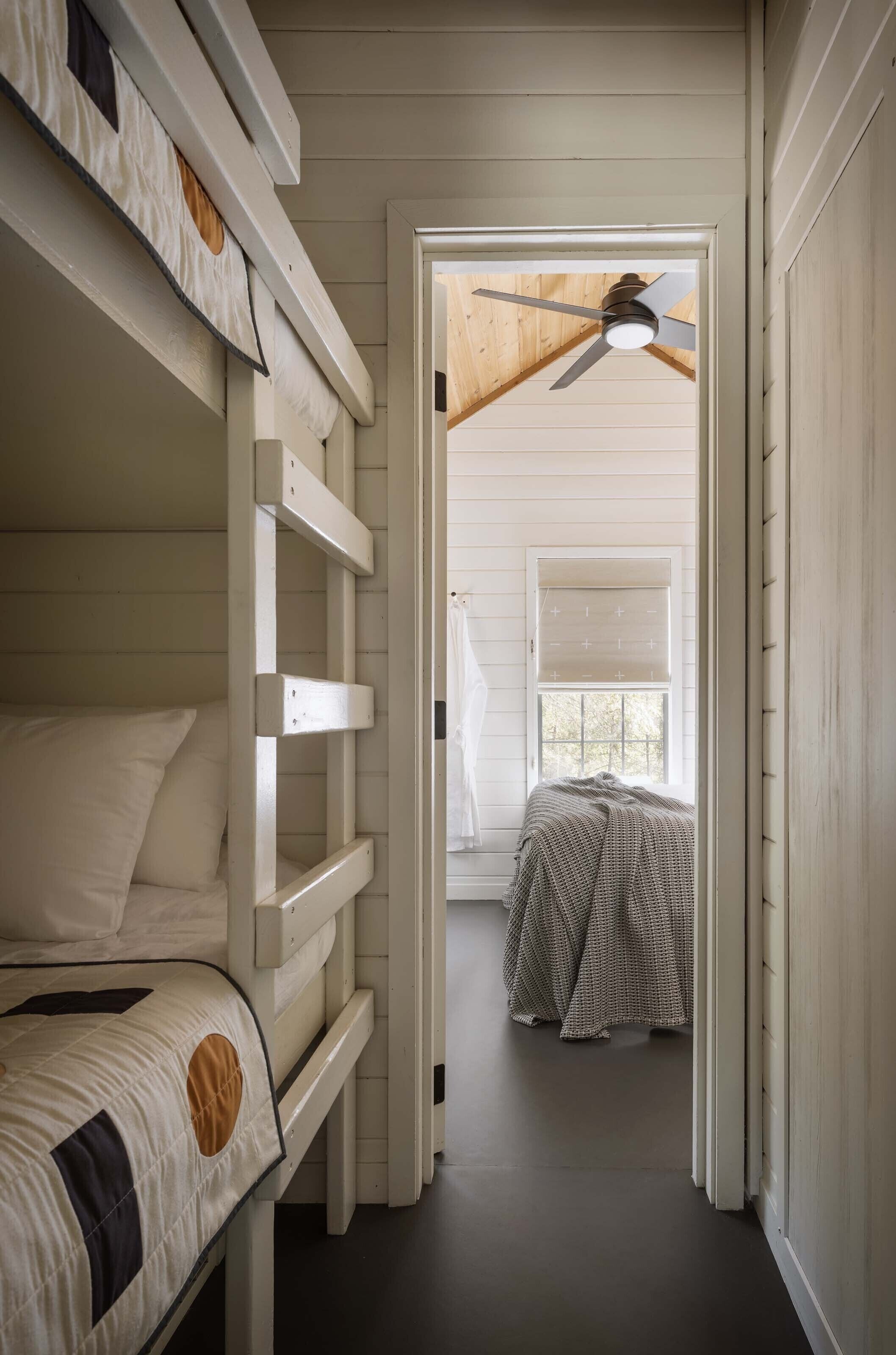 A room with bunk beds, a ceiling fan, a window with blinds, and a bed with a patterned blanket.