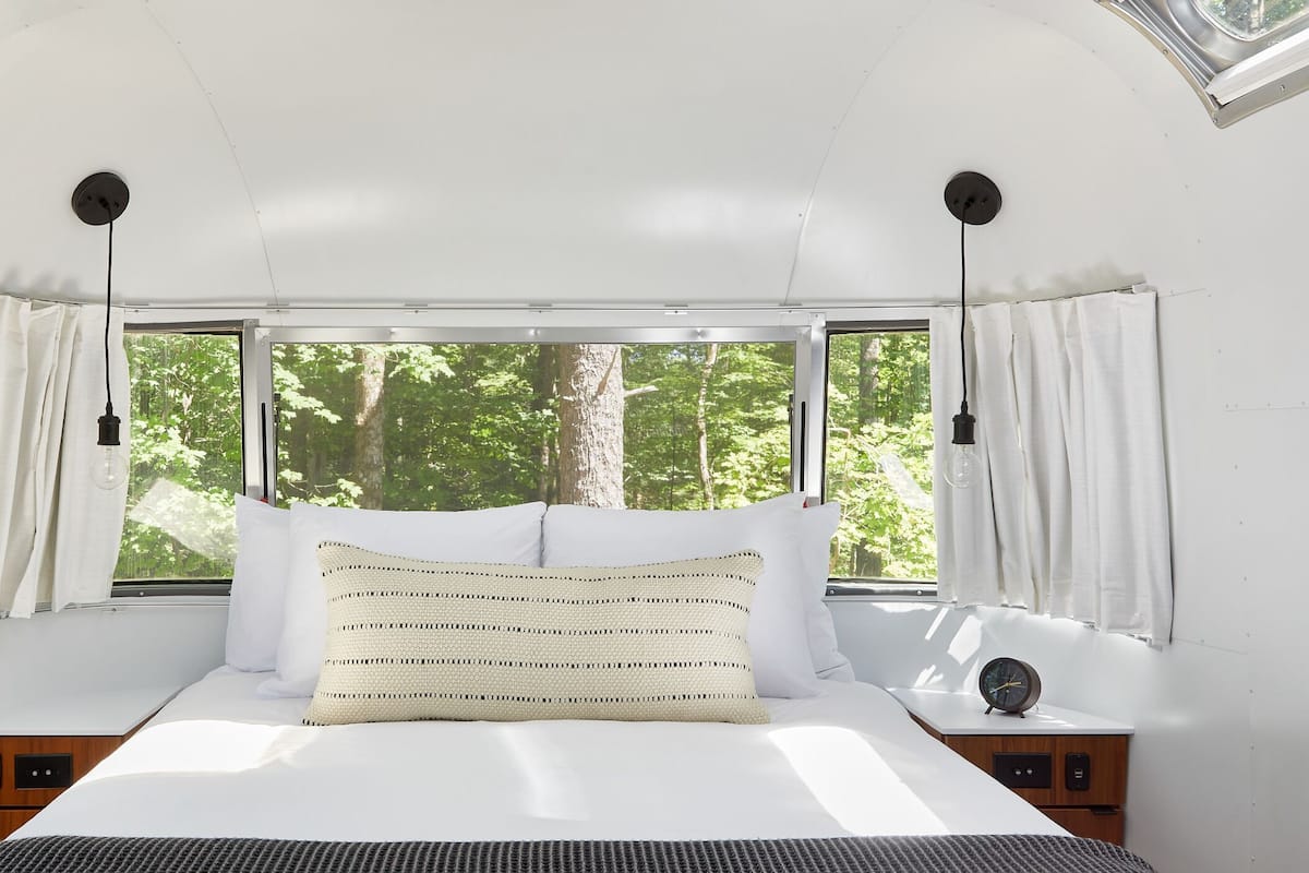 A bed with white bedding, a woven pillow, and a bedside table with a clock.