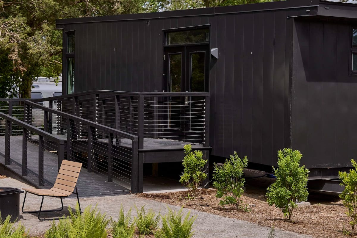 A modern building with a black exterior, a wooden deck with a metal railing, and a small outdoor seating area with a bench and a trash can.