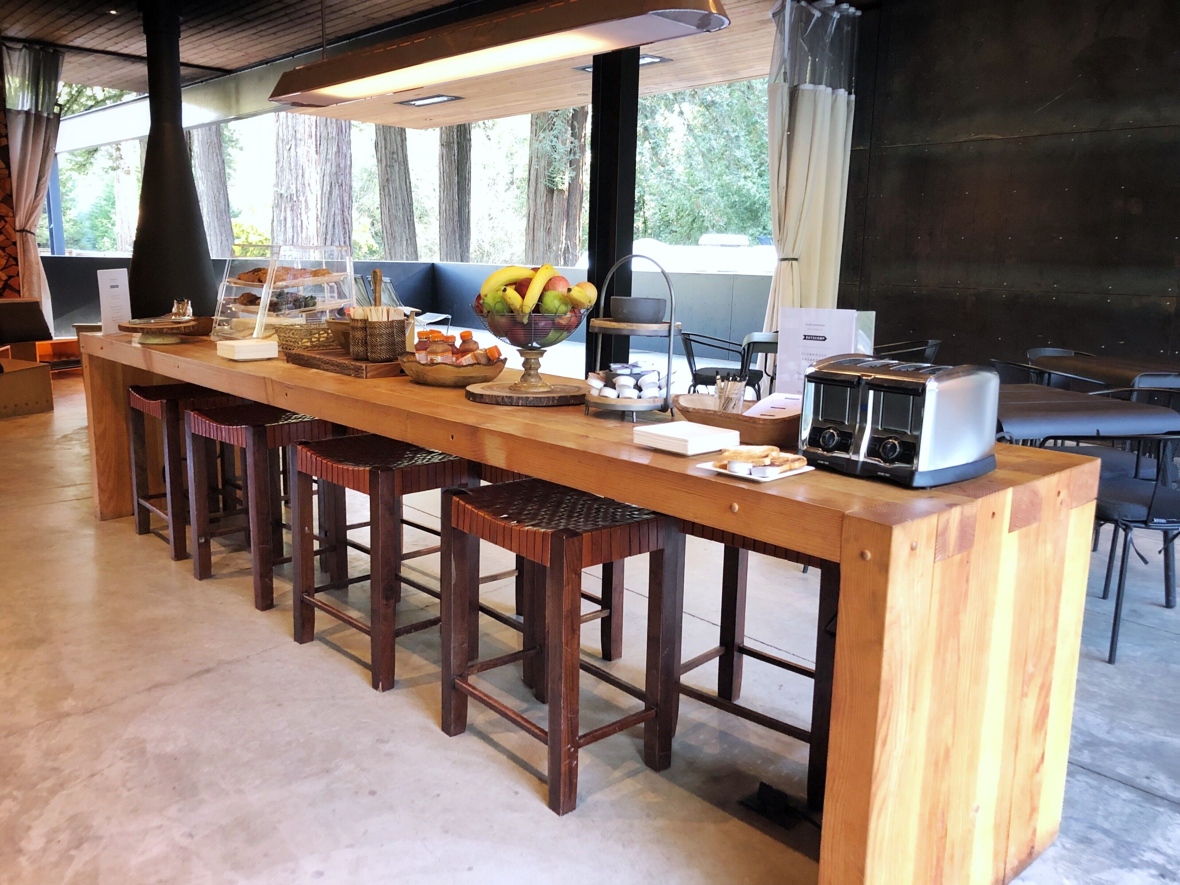 Un long comptoir en bois avec des tabourets, un panier de fruits, un grille-pain et une vue sur les arbres à l’extérieur.