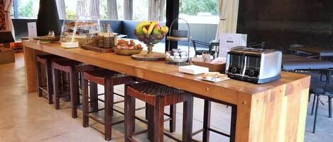 Un long comptoir en bois avec des tabourets, un panier de fruits, un grille-pain et une vue sur les arbres à l’extérieur.