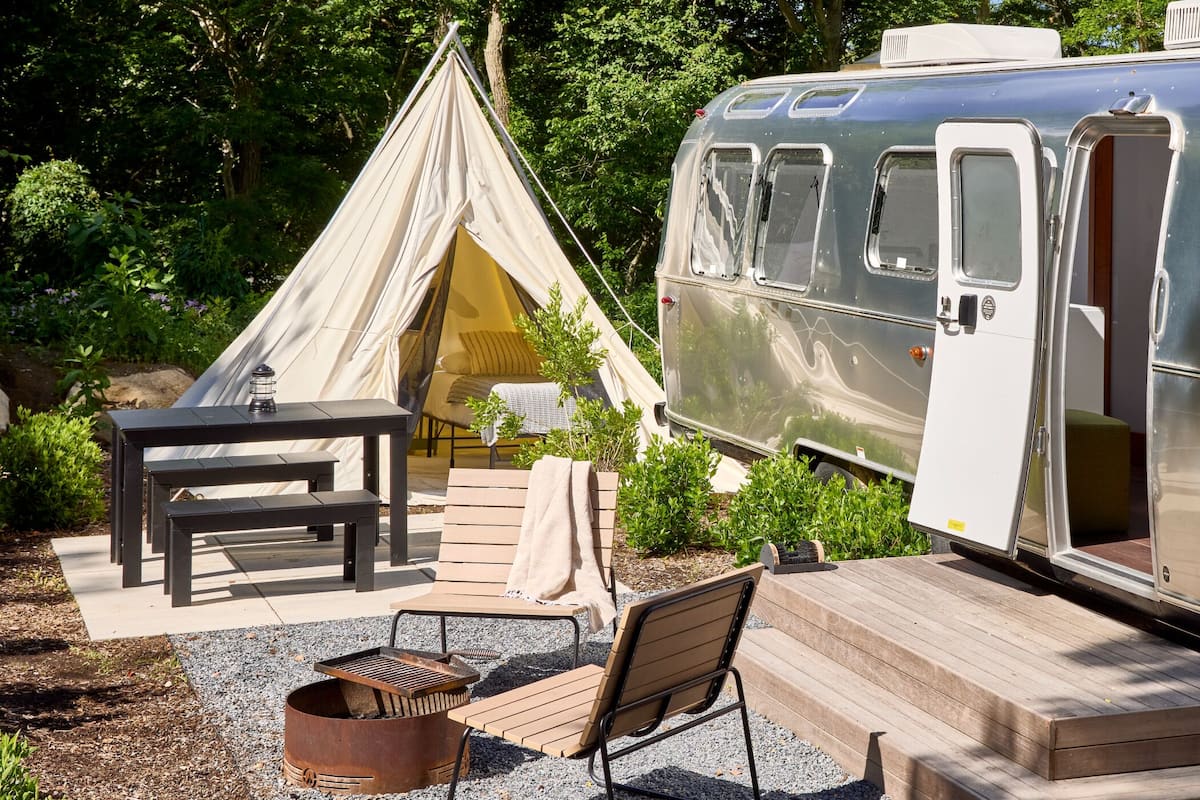 A silver RV with a sliding door, a tent, a table, and outdoor seating.