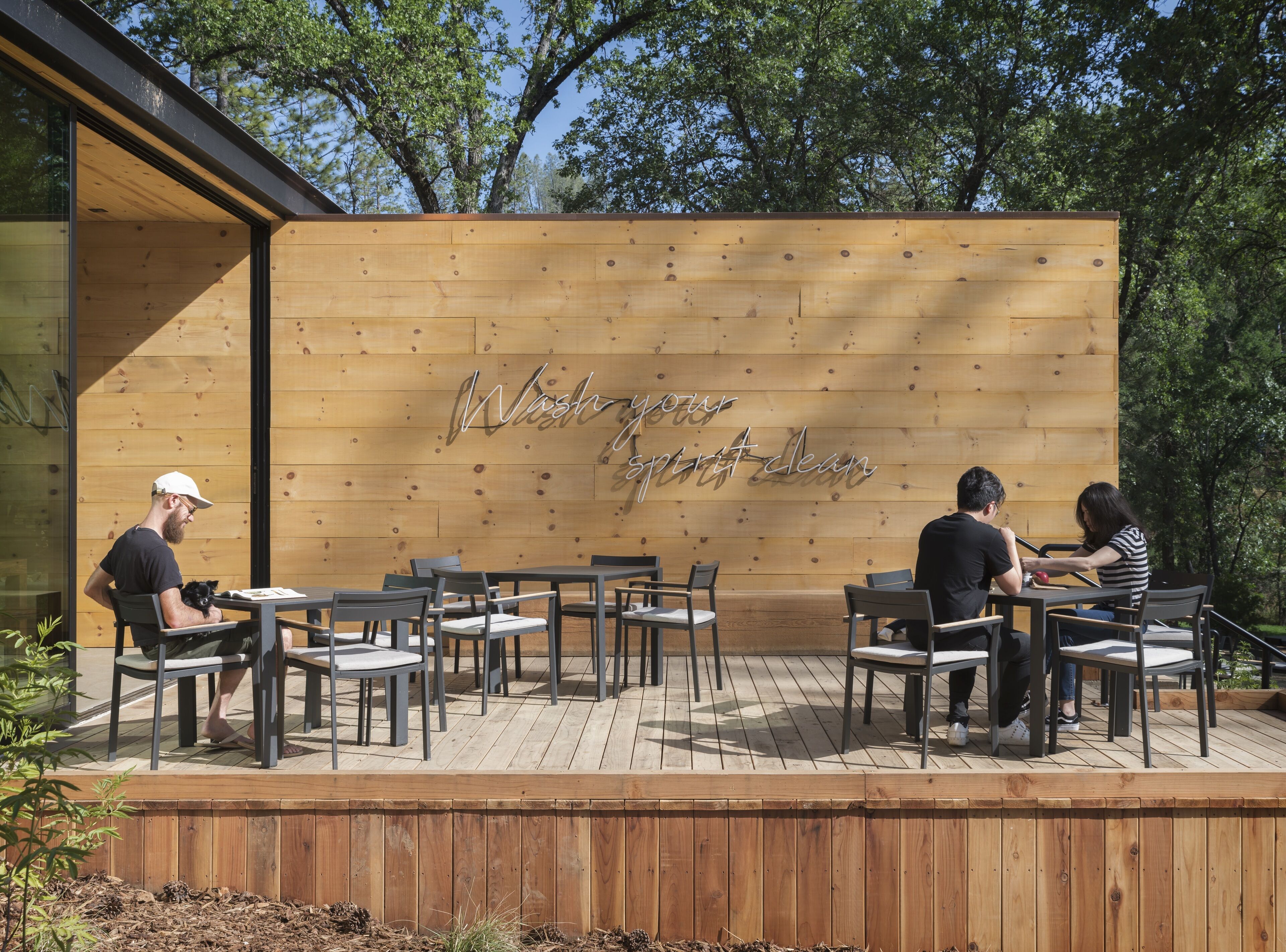 An outdoor seating area with wooden tables and chairs, a wooden wall with a message, and trees in the background.