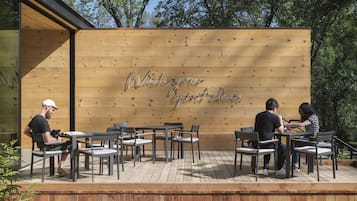 An outdoor seating area with wooden tables and chairs, a wooden wall with a message, and trees in the background.