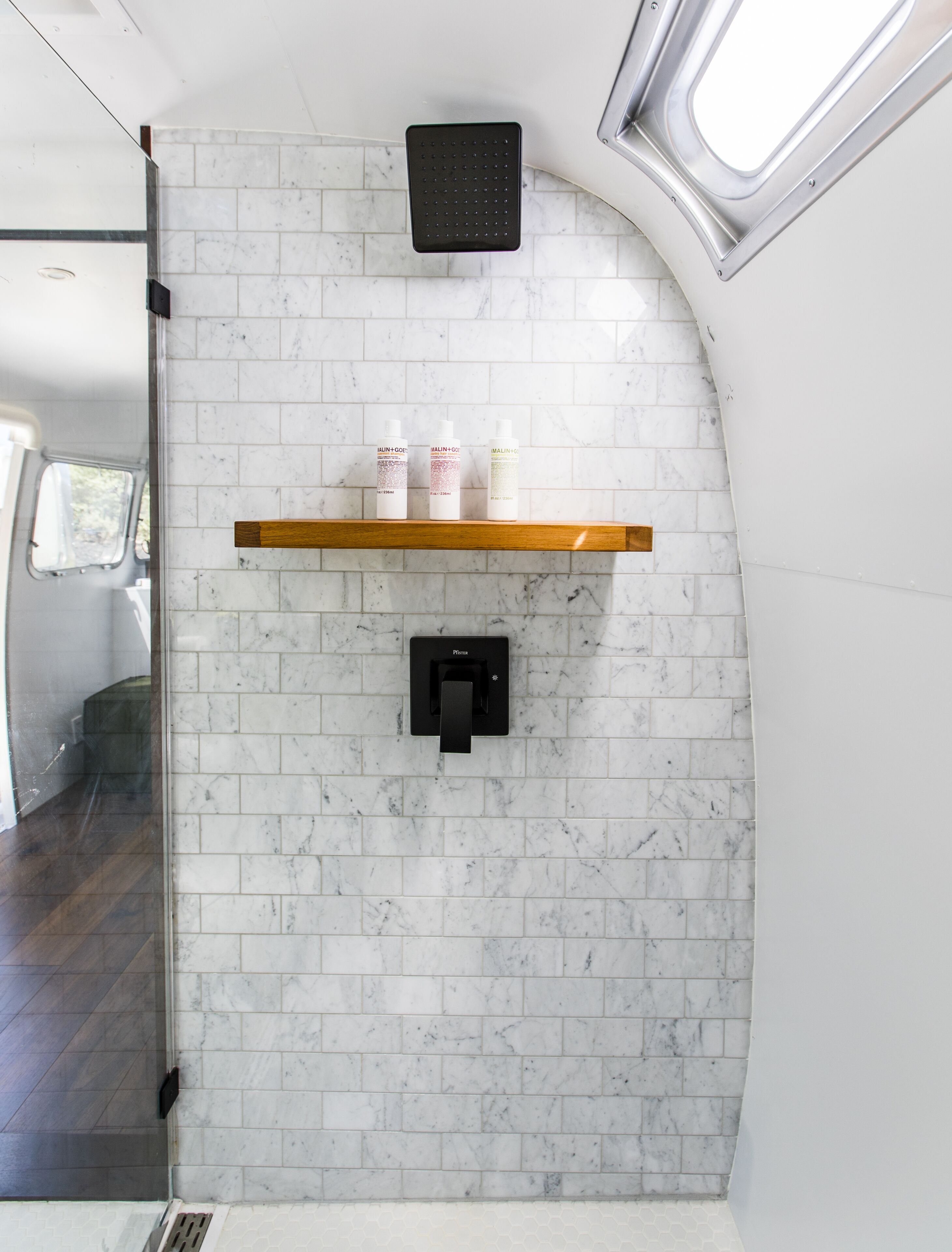 A bathroom with white marble tiles, a black towel dispenser, and three bottles on a shelf.