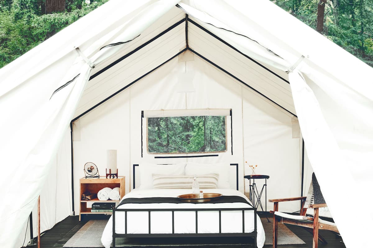 A tented accommodation with a bed, bedside table, and a view of the forest.