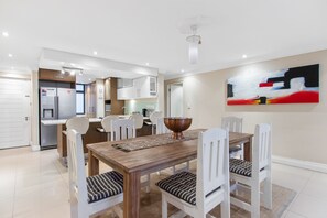 Un comedor moderno con una mesa de madera y sillas blancas, una cocina al fondo y una obra de arte contemporánea en la pared.