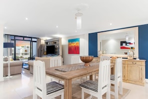 A modern living room with a wooden dining table, white chairs, a large window with a city view, a flat-screen TV, and a red abstract painting. - Eden on the Bay 108 (Cape Town)