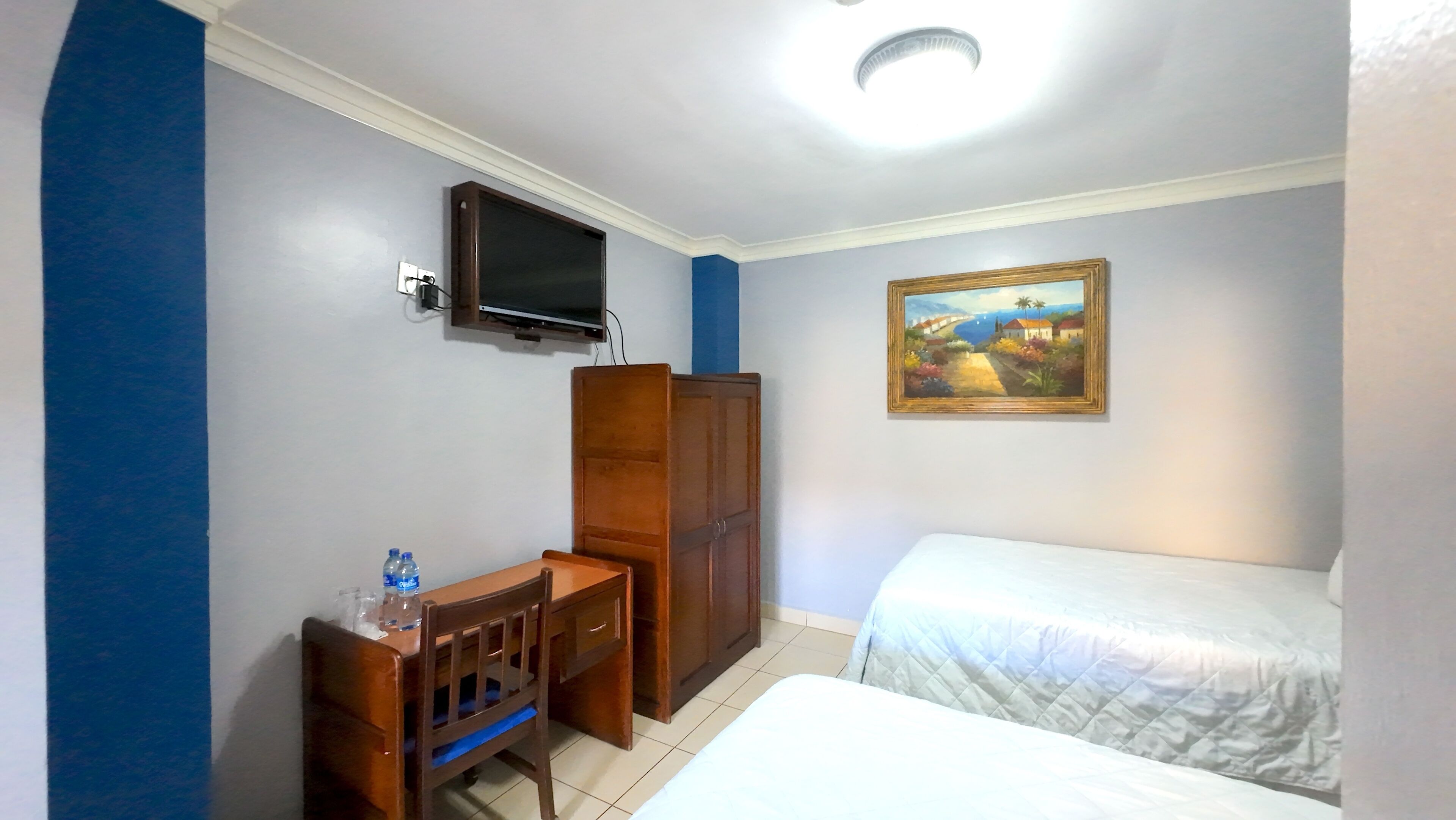 Ein Hotelzimmer mit Bett, Schreibtisch mit Stuhl, Kleiderschrank, Wandfernseher und einem Wandbild.