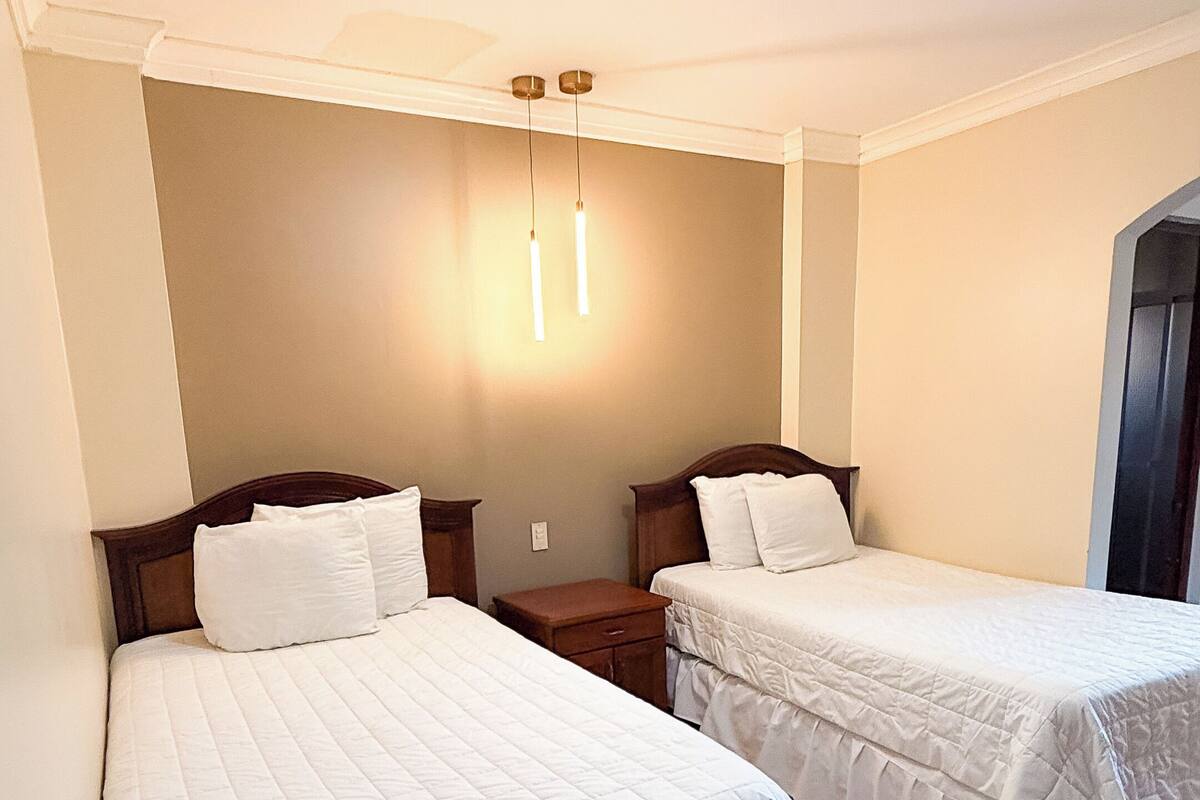 Habitación de hotel con dos camas, una mesita de noche, una lámpara de pared y una lámpara en el techo.