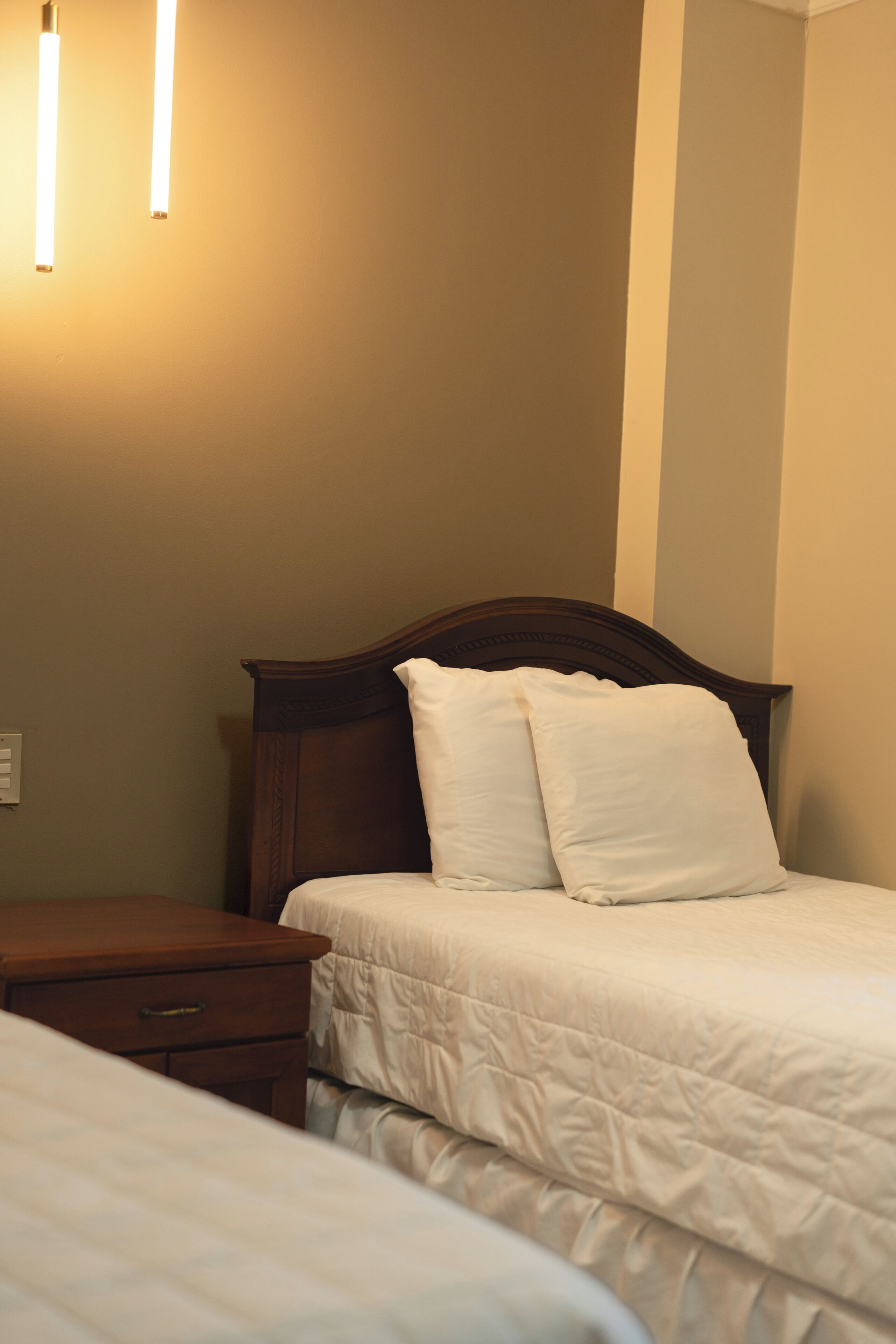 A neatly made bed with white bedding and two pillows, a wooden nightstand with a drawer, and a wall-mounted light fixture.