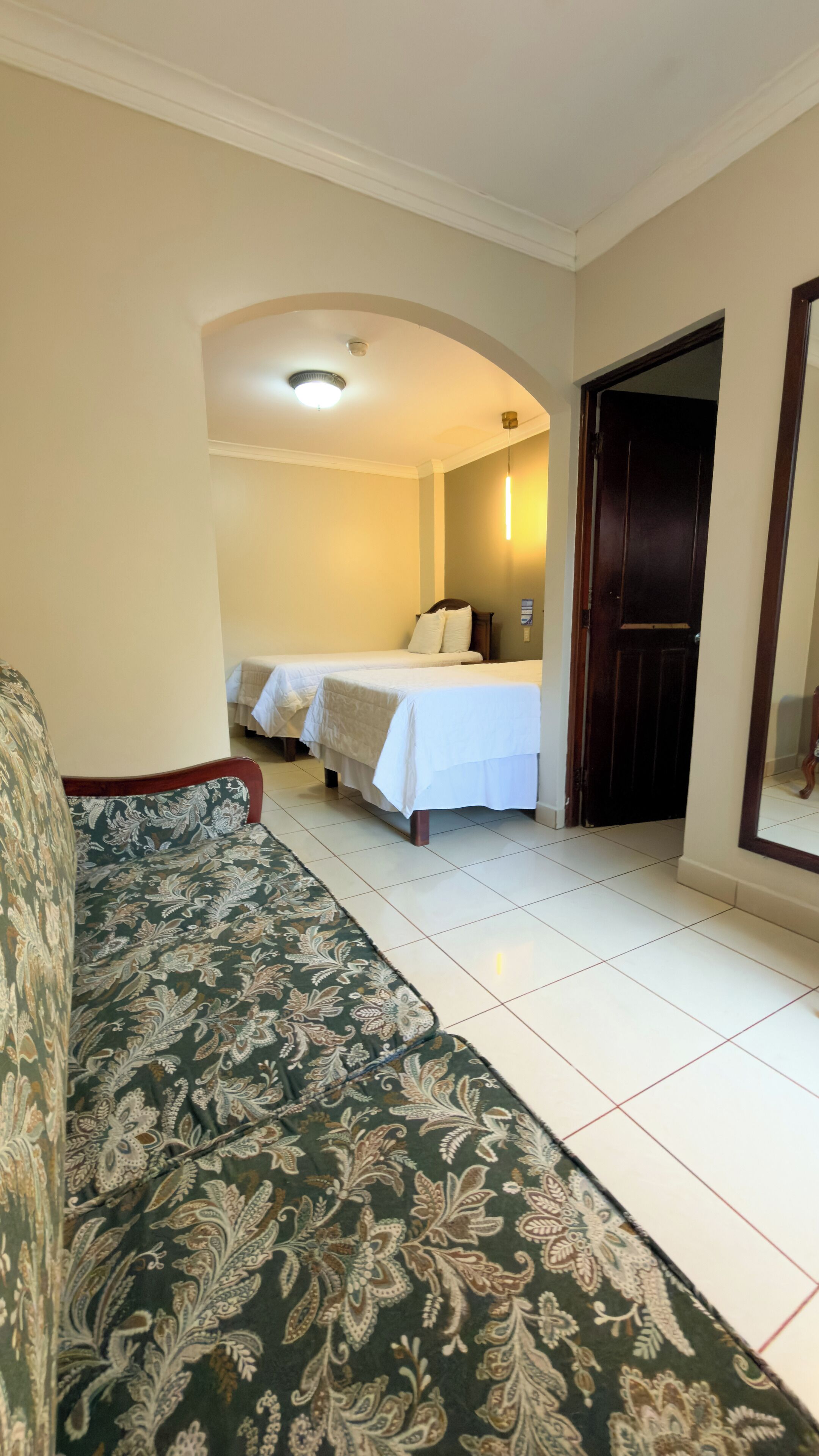 A hotel room with a green patterned sofa, two single beds with white bedding, a wooden door, and a tiled floor.