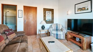 A living room with a brown sofa, a patterned armchair, a coffee table, a TV on a wooden stand, and a framed picture on the wall. - Apartment Kornelius (Carvoeiro)