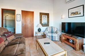 A living room with a brown sofa, a patterned armchair, a coffee table, a TV on a wooden stand, and a framed picture on the wall.