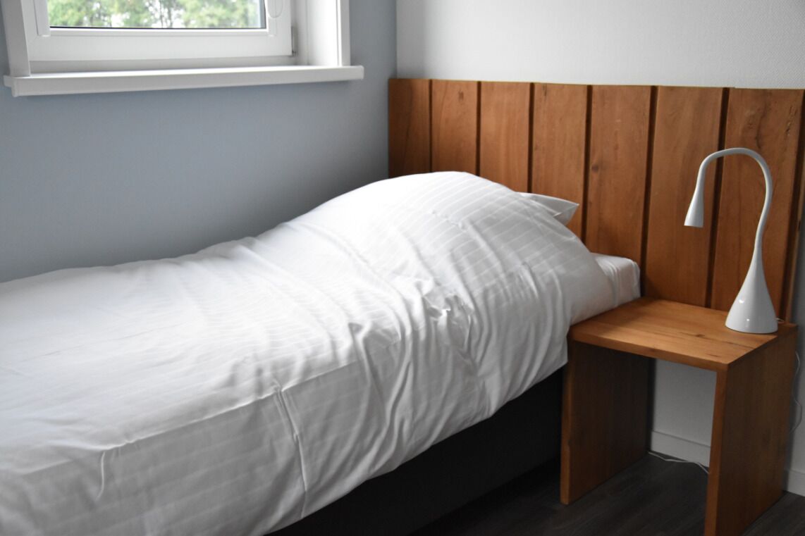 Een enkel bed met een houten hoofdeinde, een nachtkastje met een lamp en een raam boven het bed.