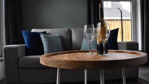 Ein modernes Wohnzimmer mit grauem Sofa, blauen und türkisen Kissen, einem Holztisch mit einer Weinflasche und zwei Gläsern sowie Blick auf eine Holzterrasse durch ein Fenster.