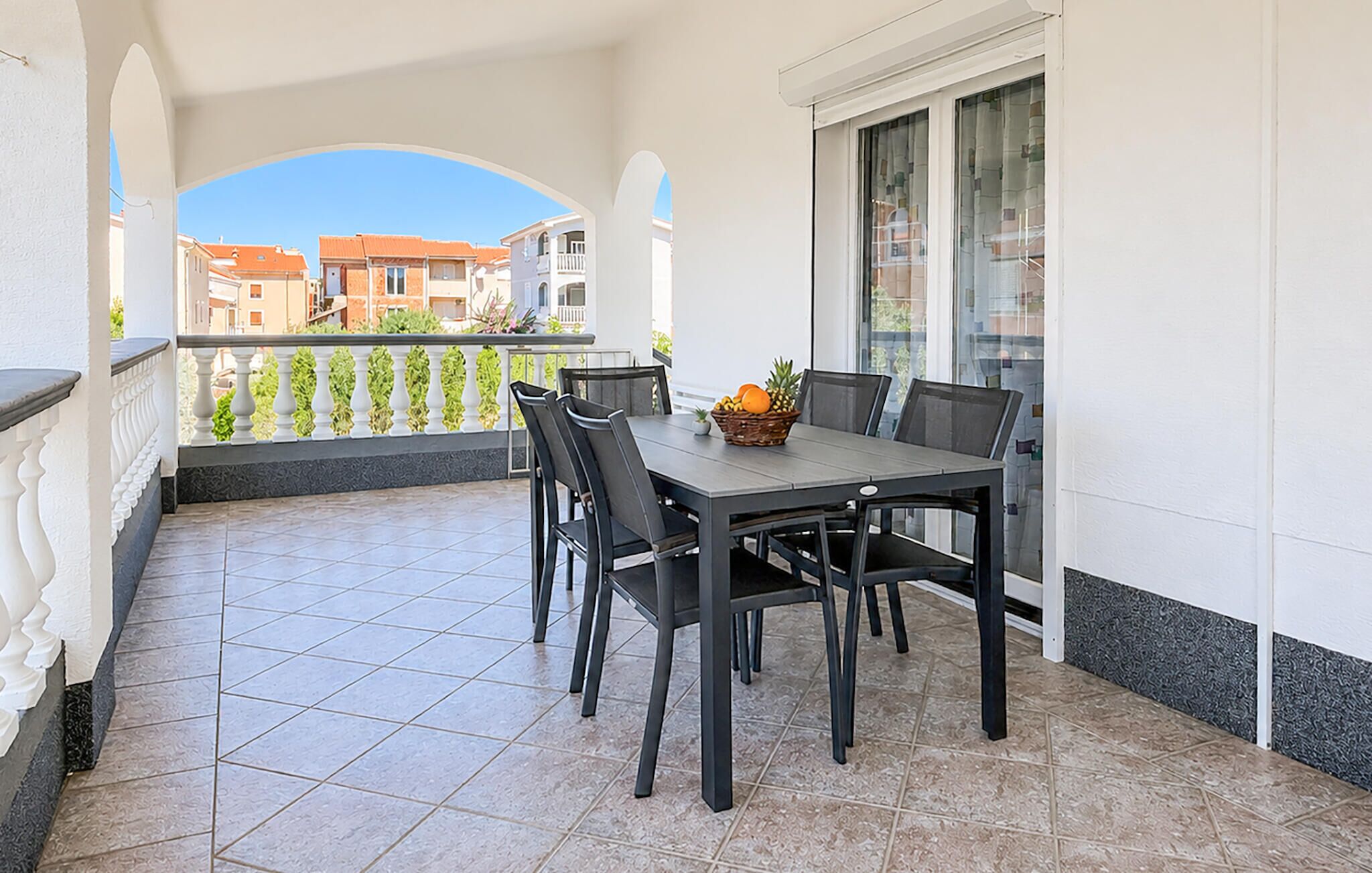A balcony with a table and chairs, a basket of fruit, and a view of buildings in the background.