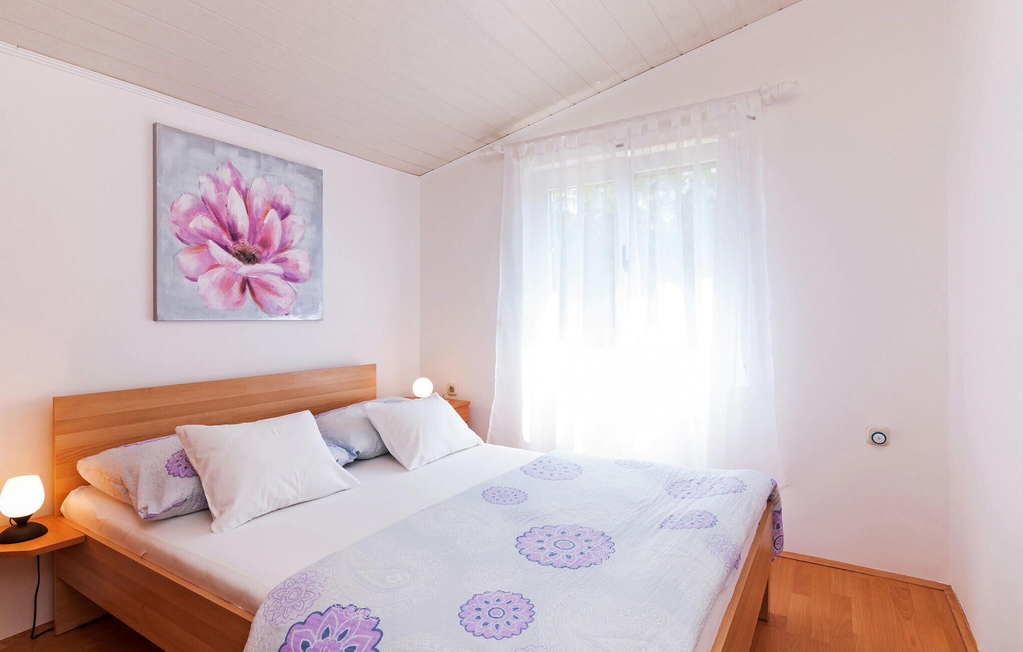 Une chambre à coucher avec un lit, une table de chevet, une lampe, une fenêtre avec des rideaux légers et un tableau floral accroché au mur.