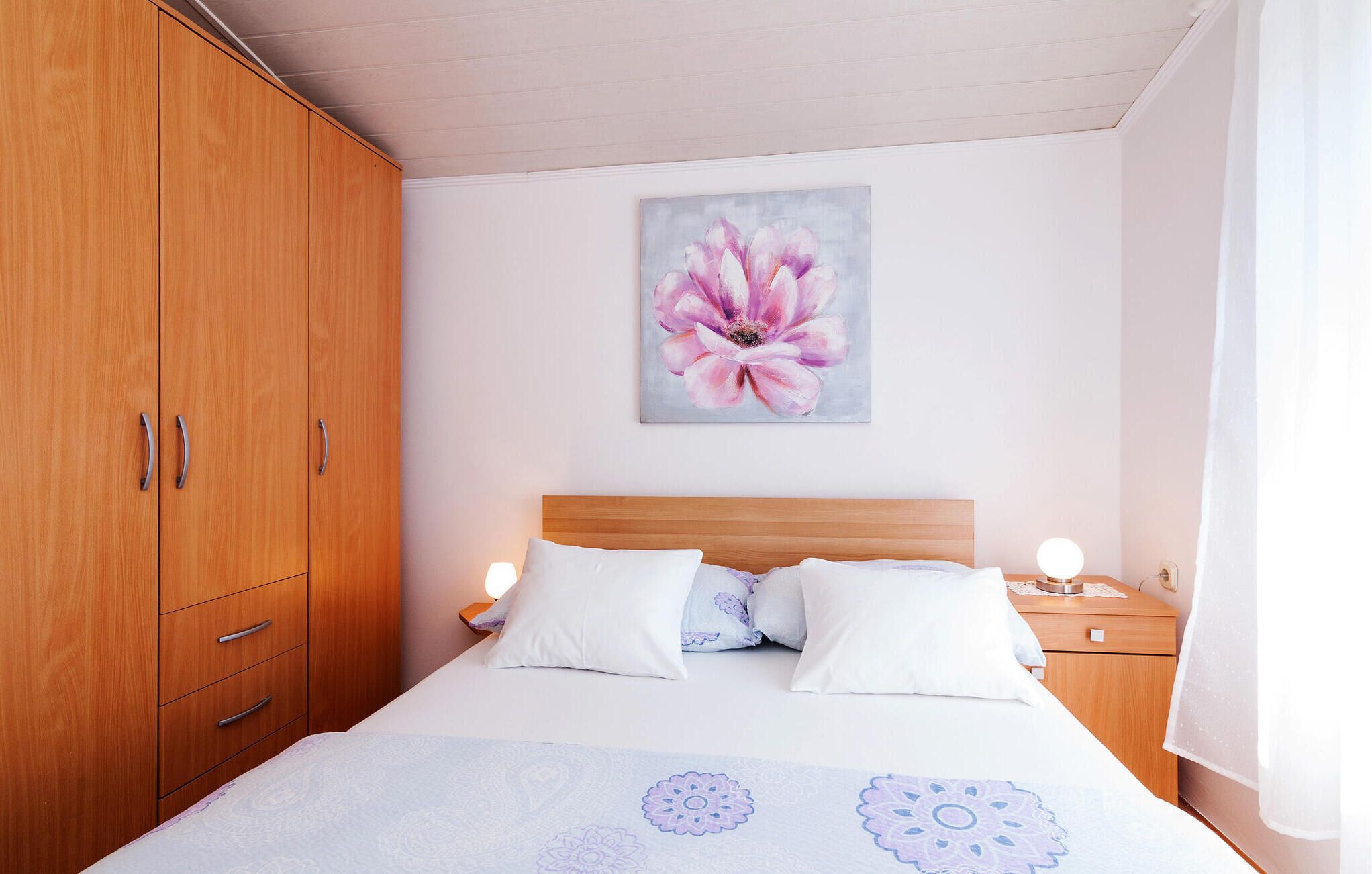 A bedroom with a wooden wardrobe, a bed with white and patterned bedding, a nightstand with a lamp, and a wall-mounted flower painting.