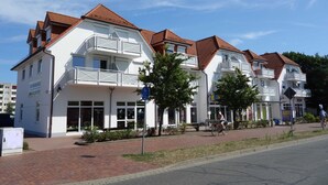 A row of white buildings with red roofs and balconies, a paved walkway, and a cyclist on the road. - Vacation object Graal Müritz A1 - F01 (Graal-Müritz)