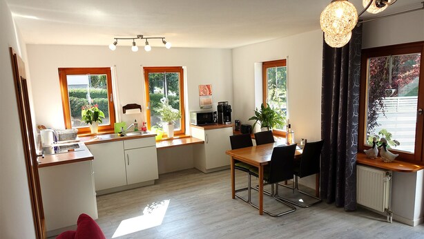 Cocina compacta con comedor, encimeras de madera y una mesa con sillas. Ventanas que ofrecen luz natural y vistas a la vegetación exterior.