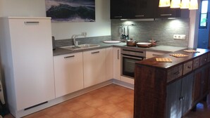 A modern kitchen with white cabinets, a stainless steel sink, and a built-in oven. There are hanging pendant lights above the counter and a painting of the ocean on the wall.