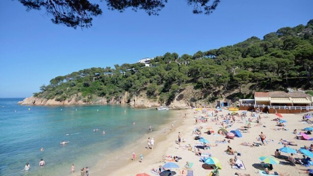 A beach with clear turquoise water, people sunbathing, and a hill with buildings in the background. - Holiday rental apartment with swimming pool in Begur, Aiguablava (Begur)