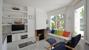 A living room with a television, shelves with model ships, a sofa with colorful cushions, and a window with a view of trees. - Holiday rental apartment with swimming pool in Begur, Aiguablava (Begur)