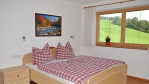 A wooden bed with red and white checkered bedding, a nightstand, a wall-mounted painting of a river, and a window with a view of a green field.