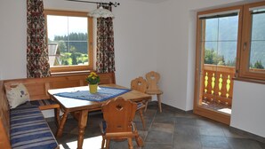 A dining area with a wooden table, chairs, and a bench. There is a window with curtains, a potted plant, and a view of greenery outside.