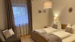 A bedroom with a wooden bed, a wooden nightstand, a bedside lamp, a sofa, and a window with sheer curtains. - Private apartment in the Dolomites: 2-8 persons (St. Lorenzen)