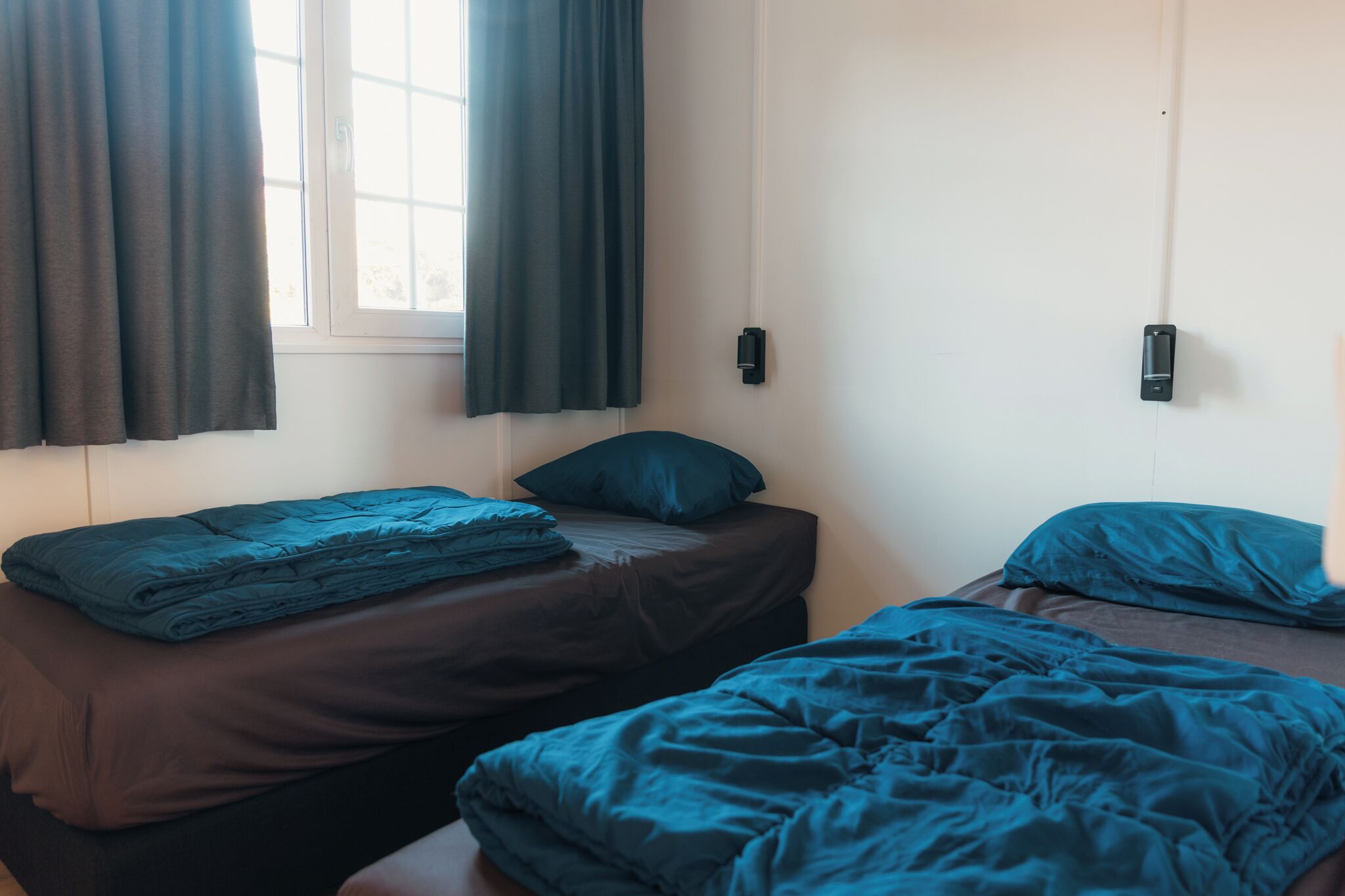 Een kamer met twee bedden, donkere beddengoed, een raam met gordijnen en een wandlamp.