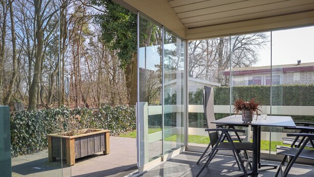 A covered patio with a table and chairs, a wooden bench, and a glass wall offering a view of trees and a hedge. - Six-person Veluwe Lounge (Voorthuizen)