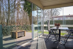 A covered patio with a table and chairs, a wooden bench, and a glass wall offering a view of trees and a hedge.