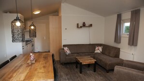 A modern living room with a wooden dining table, a brown sofa, and a small coffee table. There are hanging light fixtures and a window with curtains. - Six-person Veluwe Lounge (Voorthuizen)