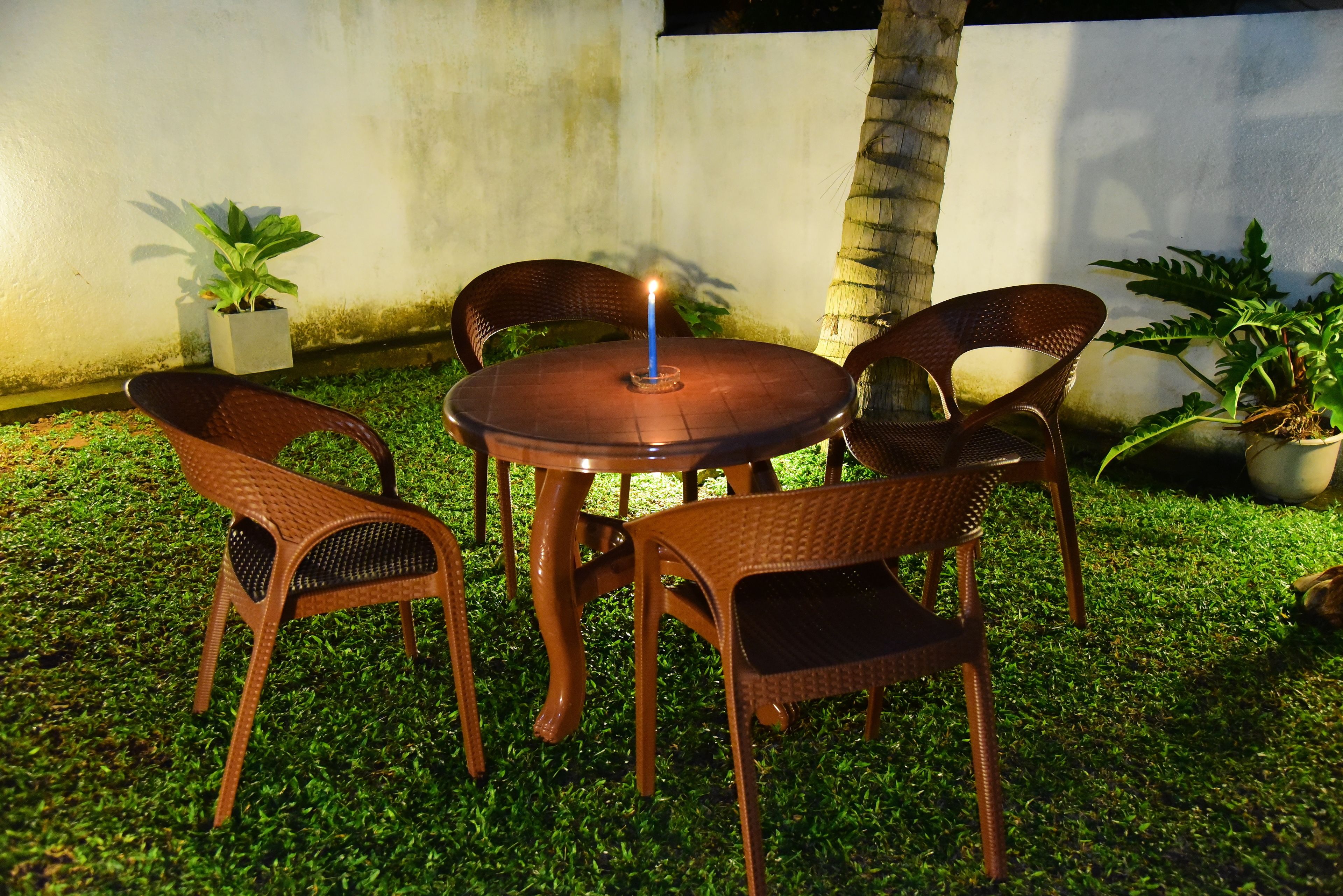 A round table with four wicker chairs, a lit candle, and potted plants in an outdoor setting.