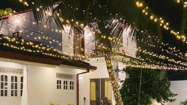 A house with a white exterior, a covered porch, and string lights. There are potted plants and a small garden area. - White Villa Airport Transit Hotel (Seeduwa - Katunayake)