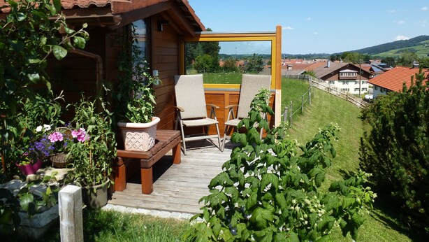 Una terraza de madera con un banco y sillas, rodeada de exuberante follaje y plantas en flor.