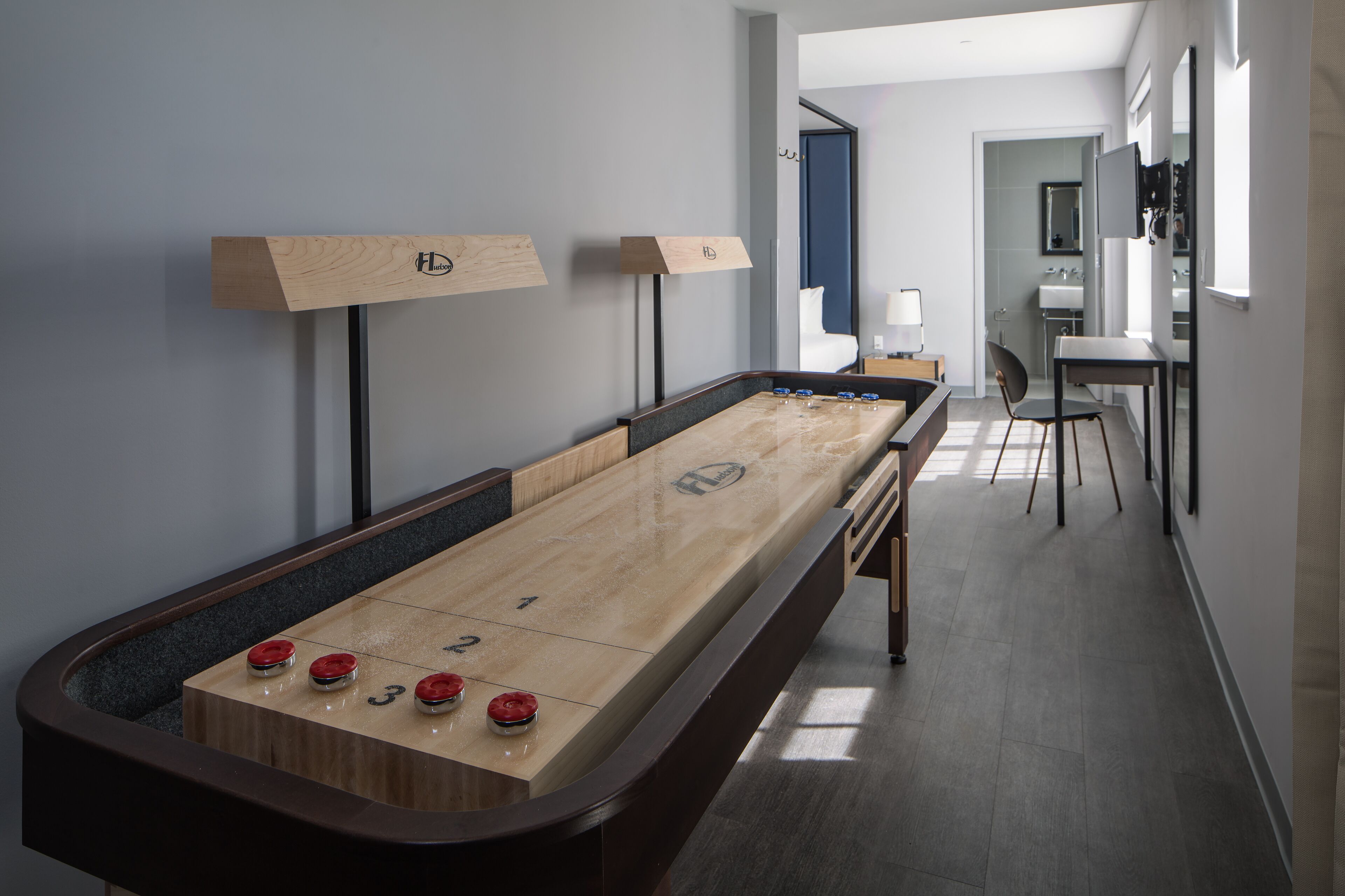 A shuffleboard table with a long, narrow layout, a small seating area with a chair, and a hallway leading to a bathroom.