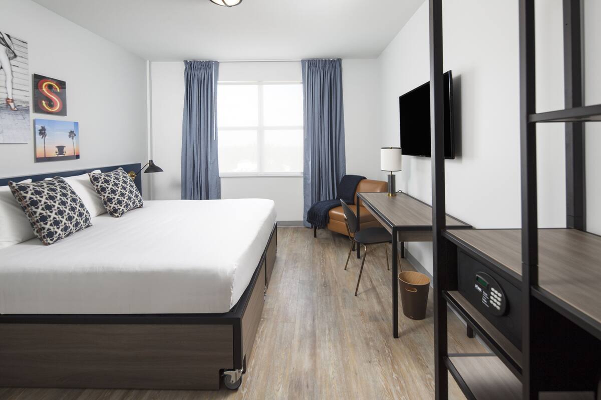 Habitació d'hotel moderna amb llit, escriptori, cadira, televisió i finestra amb cortines.