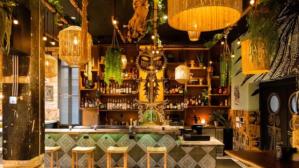 Una zona de bar con piso estampado, taburetes de madera, luces colgantes y estantes llenos de botellas.