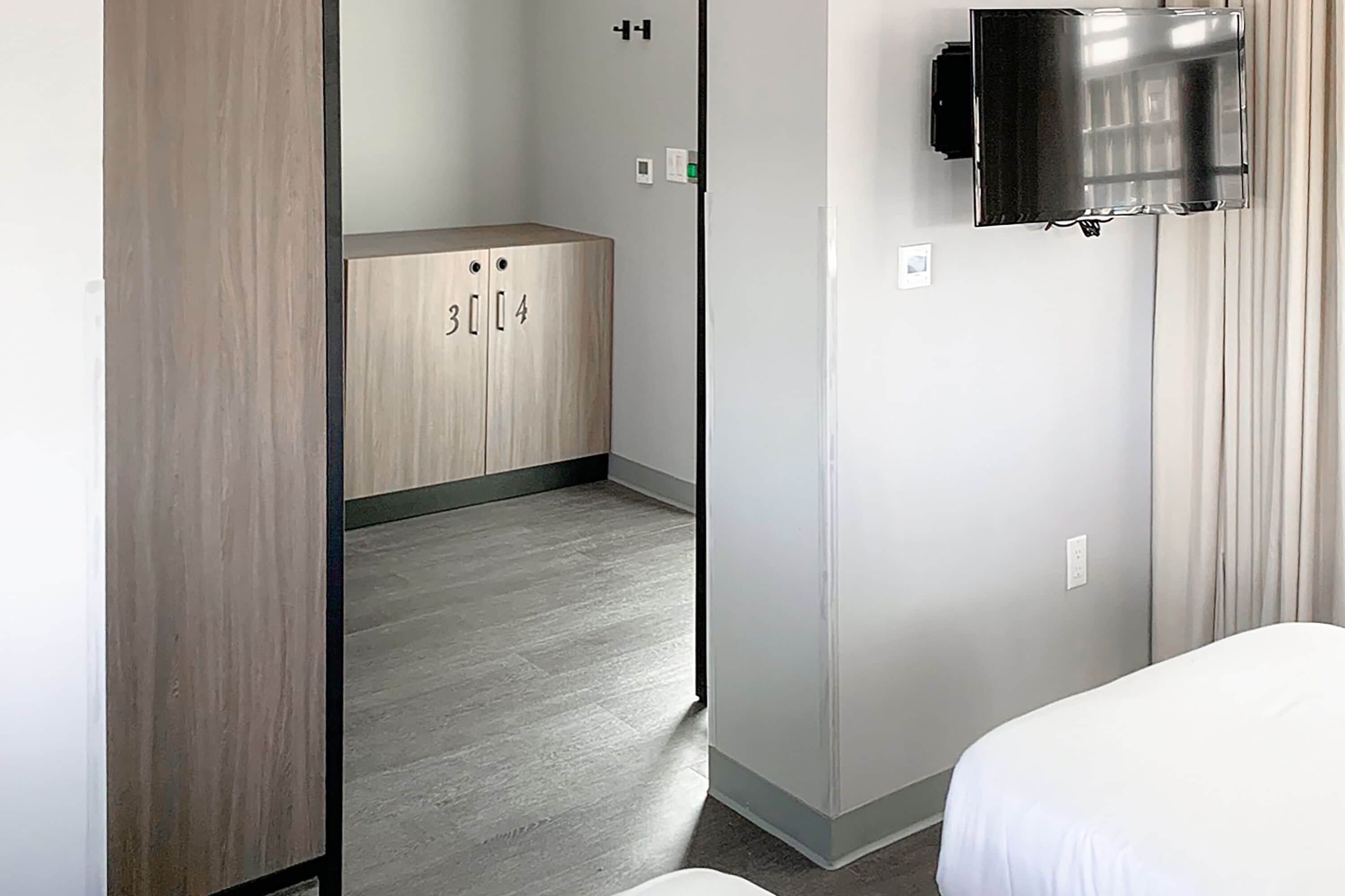 A hotel room with a bed, a TV mounted on the wall, a wooden wardrobe with numbers, and a bathroom visible through an open door.