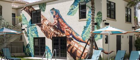 Zwembad met ligbedden en parasols, muurschildering van een dinosaurus, en een gebouw met meerdere ramen en deuren.