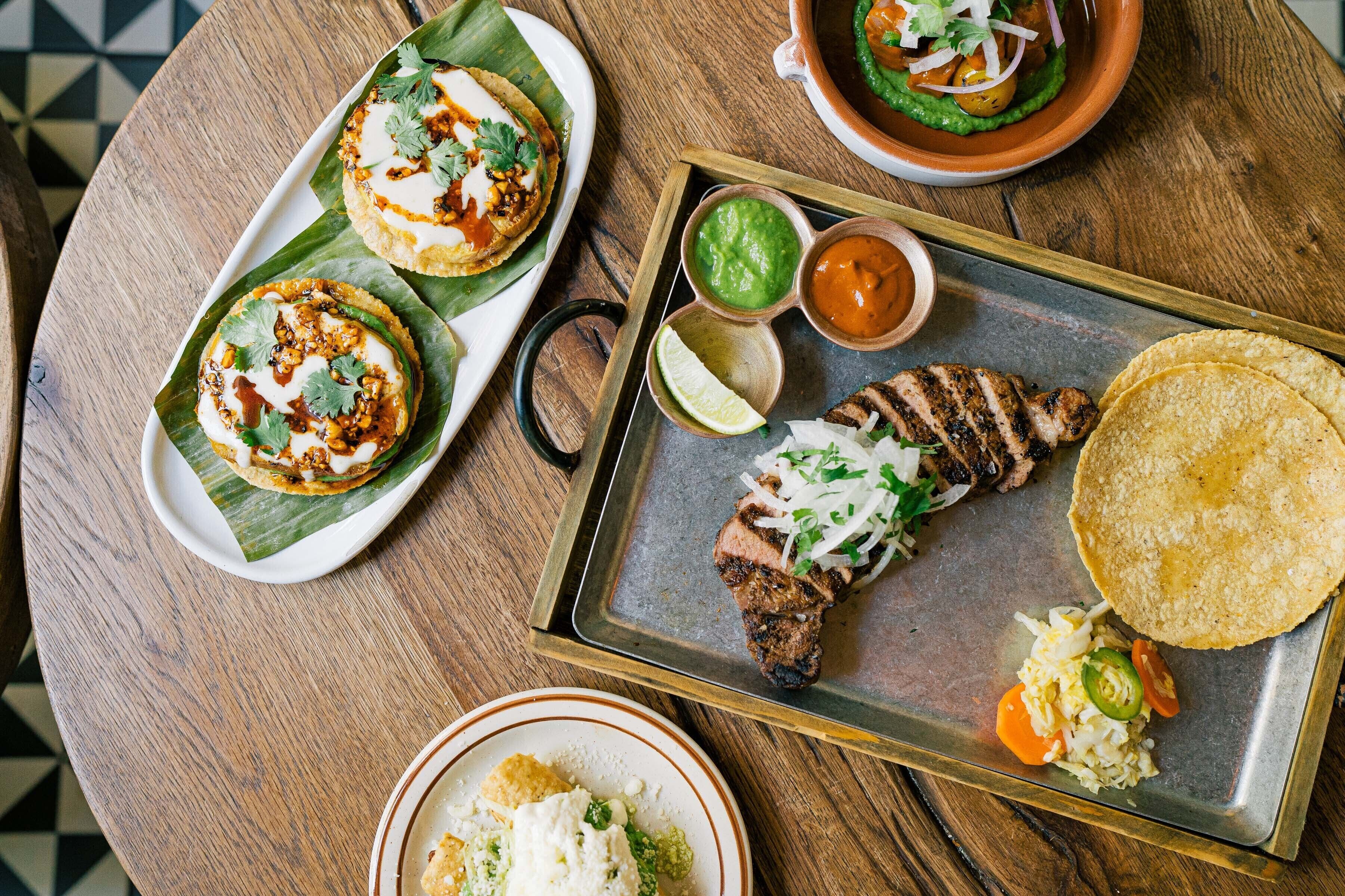 A wooden table with a variety of dishes including tacos, grilled meat, and a tortilla.