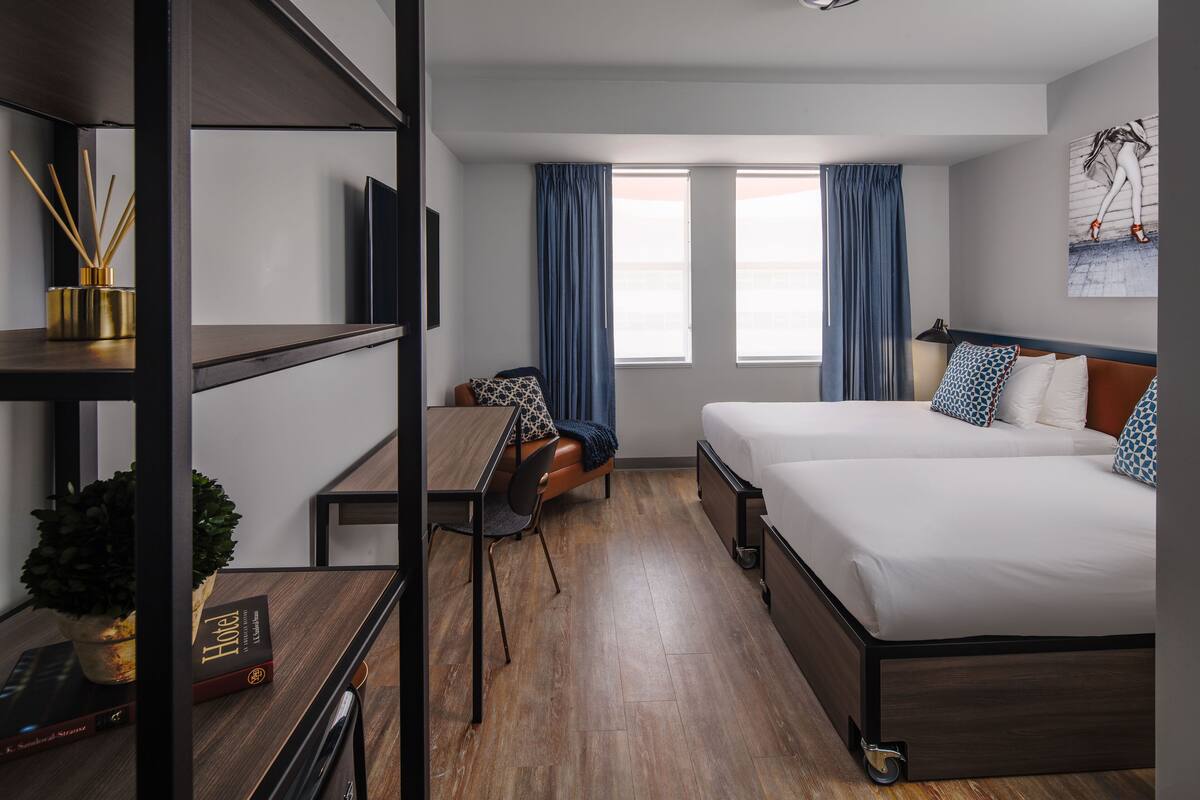 Habitació d'hotel moderna amb terra de fusta, escriptori amb cadira, llit amb almoinos decoratius i una finestra gran amb cortines.