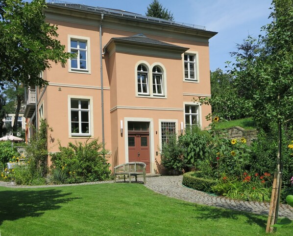 A two-story house with a front door, a small porch, and a garden with various plants and flowers. - Spacious vacation apartment on the Elbe slope - directly on the Blue Wonder (Dresden)