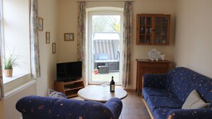 A living room with a sofa, a television, a small table with a bottle and glass, and a view through a glass door to an outdoor area. - Vacation apartment Ostseeanleger - OSTSEE-Ferienwohnungen (Hasselberg)