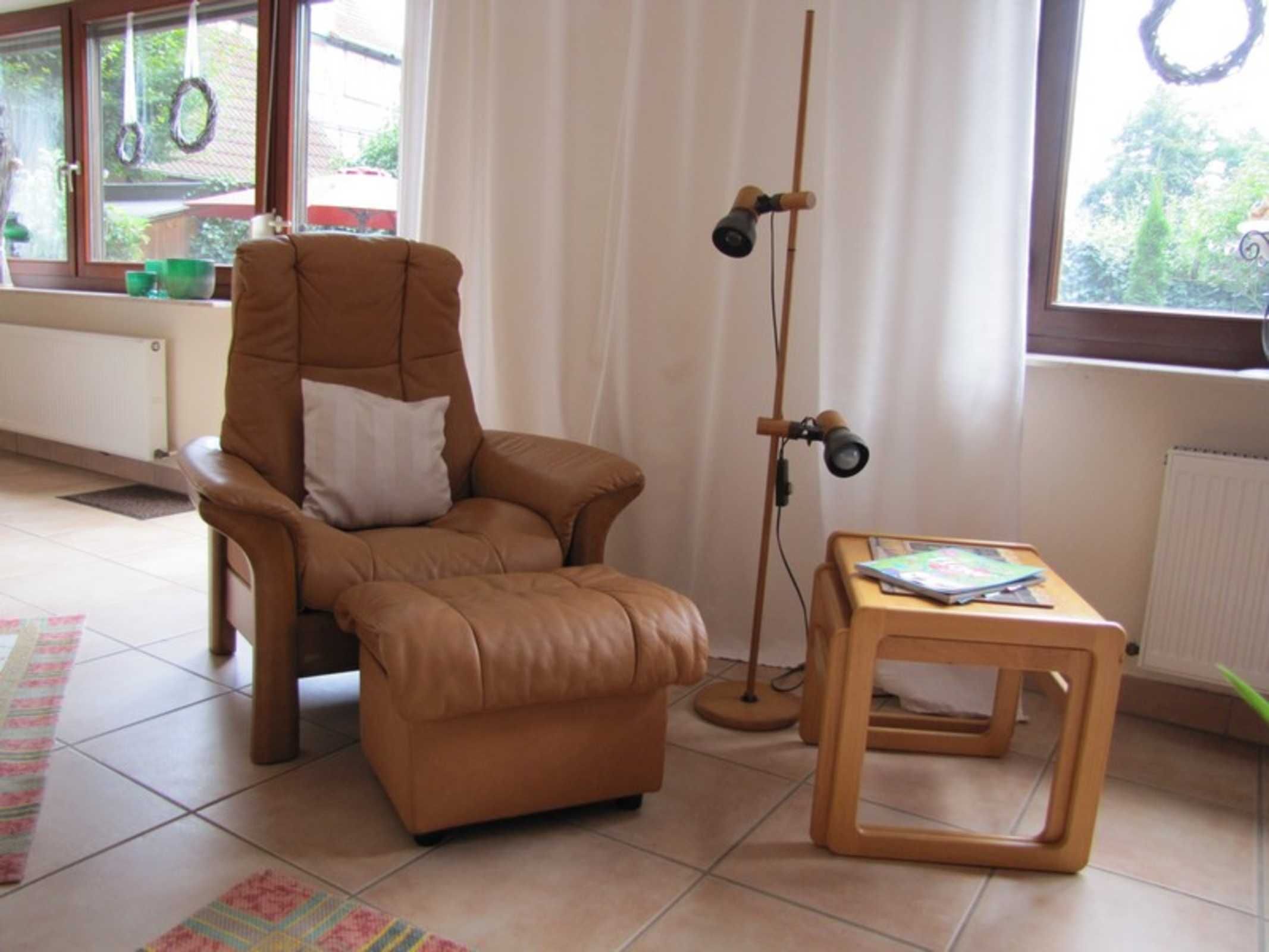 Ein Zimmer mit braunem Ledersessel, einem Holztischchen, einer Stehlampe und Blick auf Grünfläche außerhalb.