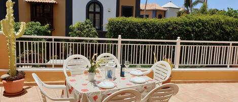A patio with a table set for two, surrounded by white chairs, with a cactus and potted plants nearby.